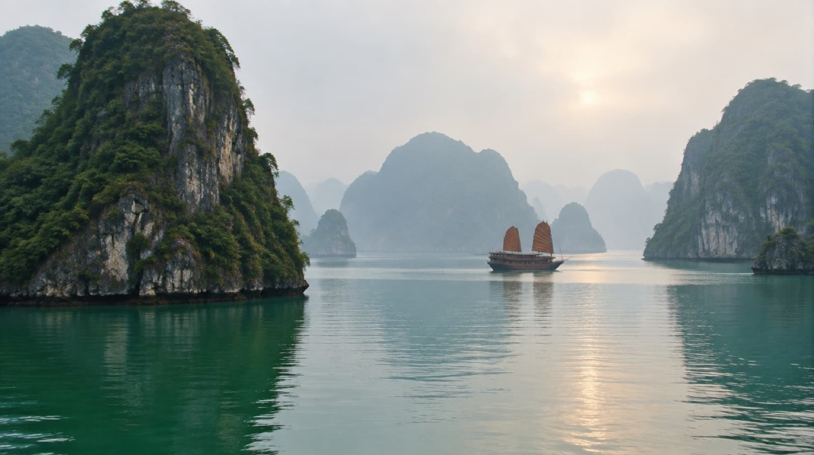 Limestone karst islands rising from calm green water in Ha Long Bay, a junk boat in the distance