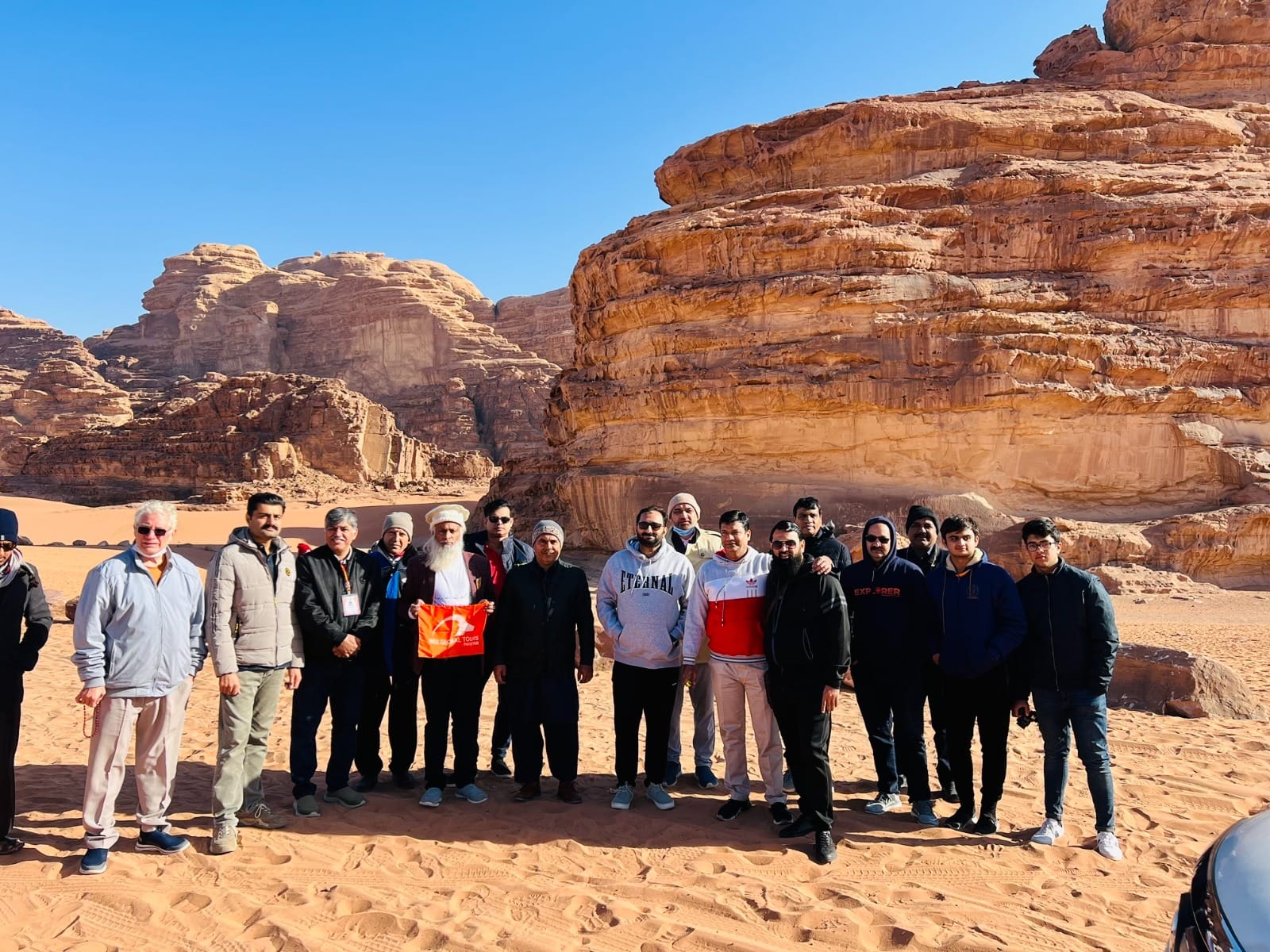 An IBEX group photographed on the red sand at Wadi Rum, sandstone cliffs behind, one traveller holding the IBEX Global Tours flag