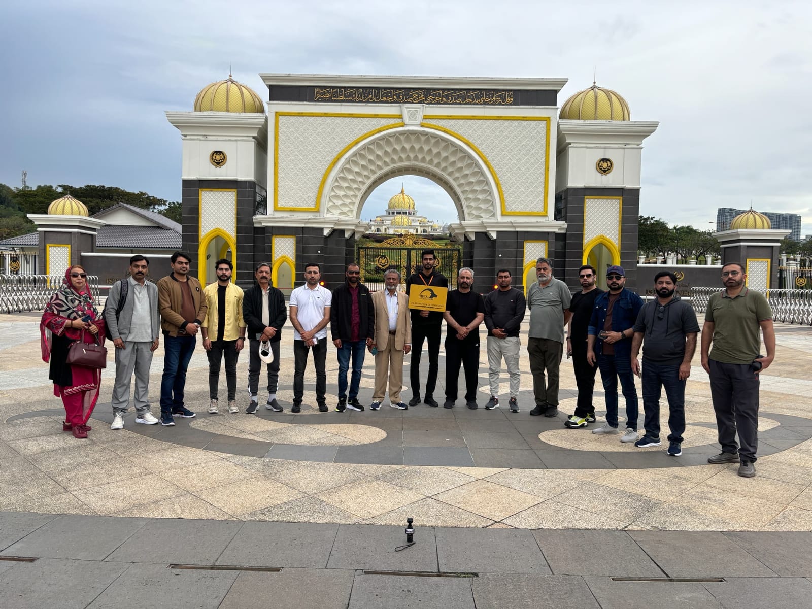 An IBEX group photographed in front of the gates of the Istana Negara in Kuala Lumpur, one traveller holding an IBEX Global Tours board